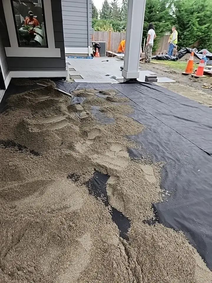 Paver patio installation in progress with aggregate spread over black geotextile fabric beside a home in Western Washington